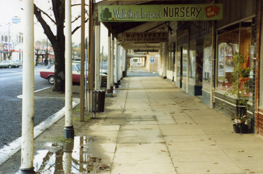 Footpath outside Tippett's store and Ross's Gift Store. Sign for Ross's shop reads 'Walk thru & Inspect Nursery. At rear." Sign for Tippett's reads "RJ Tipett Drapery Furniture".