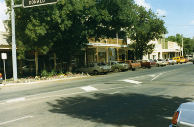 Shops at west end and south-side of High Street. 'Donald' on road sign in top left of photo. Large tree in garden bed in front of shops. Sign on shop "Ross's Gifts'.