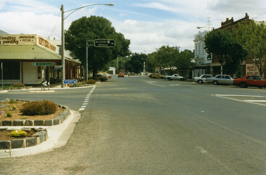 Garden beds in lower LH, road signs pointing to St Arnaud and to Mildura and Donald. North Central Panel Beaters on corner. Orange car driving west.