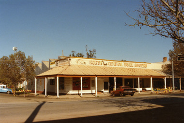 White brick building with varanda.  Red car in front of shop on High St, blue & white vehicle on LH side on Armstrong St. 