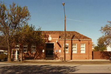 Charlton Post Office. Red brick single storey building. Clock above entrance shows 5 minutes to nine. Yellow combi van, tree and public telephone in front.
