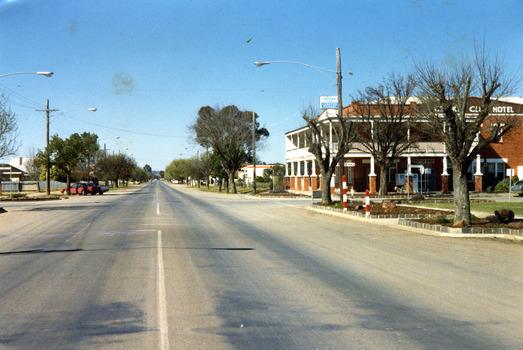  Two storey, red brick Cricket Club Hotel on RH side of photo. Red car and white ute parked on the other side of the street. Red and white school crossing posts in garden bed on RH side.