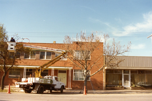 Two storey red brick building. Signage on the windows reads SOIL CONSERVATION AUTHORITY. A man on a cherry picker in front of building trimming a tree.