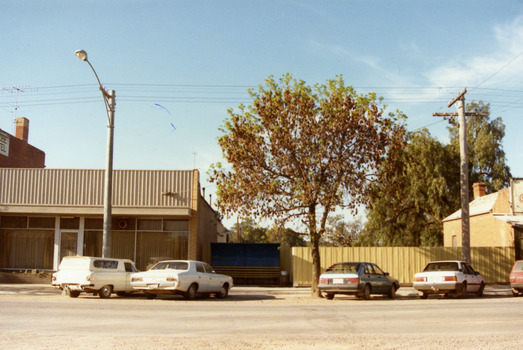 A single storey, glass fronted building with a white-framed front door.  There is a large light pole and a white panel van and a white car are in front of the building. There is a blue awning over a yellow seat to the right of the building.