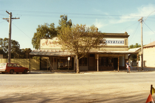Three single-storey shops with corrugated tin verandahs. Signage reads Arundell's SHOE STORE and ESTATES. Tree in front of the buildings. A red station wagon in front and a boy on a bicycle on the RH side. 