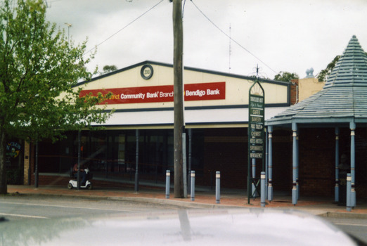 Bendigo Bank building. Single storey brick building. Signage reads 2003 Charlton & District Community Bank Branch Bendigo Bank. Blue timber covered entrance to Traveller's Rest complex. Person on mobility scooter.