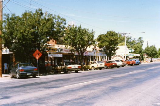 Row of shops. Cars are parked in front of the shops and there are three trees. An orange school crossing sign is next to dark blue car.