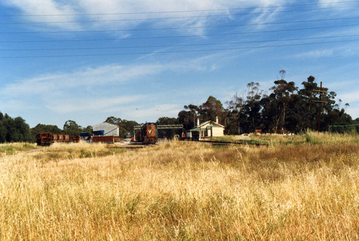 Railway Station. On the right, there is a small cream and green weatherboard building with a large verandah. There is a train engine and trucks in the middle of the photo.