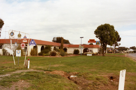 The Motel is a white brick, single-storey building with a red trimmed roof. The verandah and some front windows are arches. There is a large garden bed with shrubs in front of the verandah.