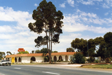 Charlton Motel taken from across the Calder Hwy looking south east. The motel is a white brick single storey building with red-brown trim on the eaves. The verandah and windows are arched.