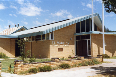 Anglican Church. White brick single storey building with an iron roof and blue fascia trim. There is a dark coloured double door with windows.