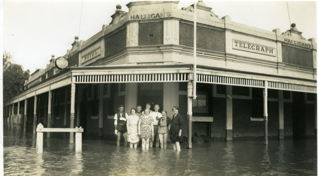 Group of eight people standing calf deep in flood water and the front of the Telegraph hotel. Sandbags line all of the entrances.