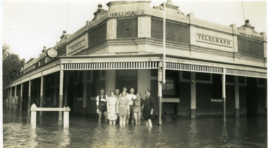 Group of eight people standing calf deep in flood water and the front of the Telegraph hotel. Sandbags line all of the entrances.