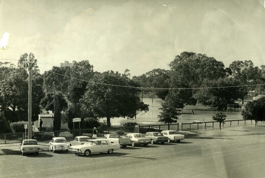 1973 flood at Gordon Park Charlton. 8 cars are lined up along road in front of park entrance. Flood waters have inundated the park.