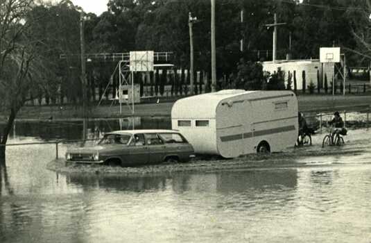 1973 flood near Gordon Park. Car and caravan driving through flood water on read followed by two bicycles.