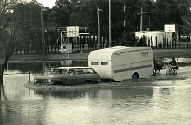 1973 flood near Gordon Park. Car and caravan driving through flood water on read followed by two bicycles.