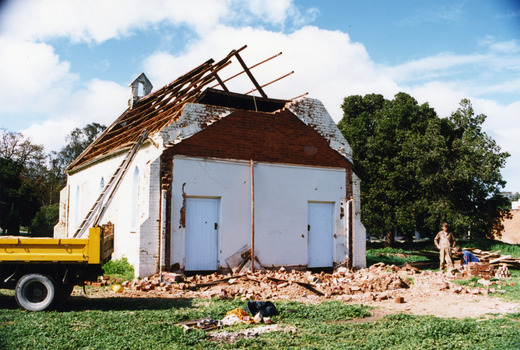 Back of St Andrews church being demolished. Brick litter the ground. Yellow dump truck in forground and a man in workwear stands to one side. Roof has been removed.