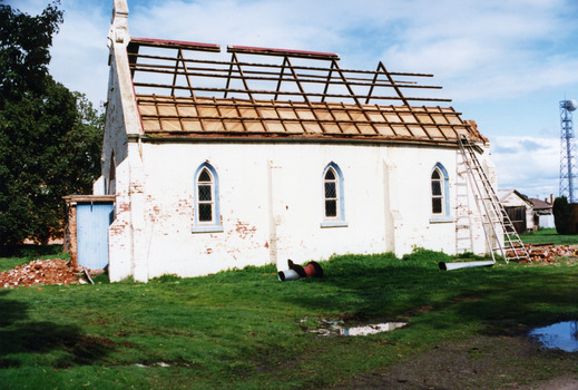 Side view of church being demolished. Roof has been removed and only timbers remain. A large ladder leans against the building. Bricks litter the ground.