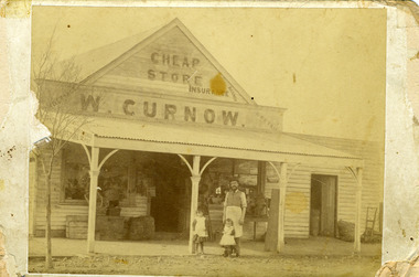 Shop keeper and two young daughters stand in front of store. Sign reads Cheap Store, W. Curnow.