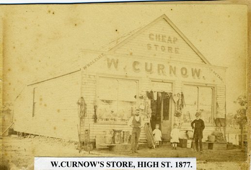 Weatherboard store with various goods hanging from front wall. Two men and two young girls stand in front.