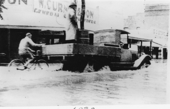 Flooded High Street with Ute driving through. A person on a bike holds onto the Ute tray.
