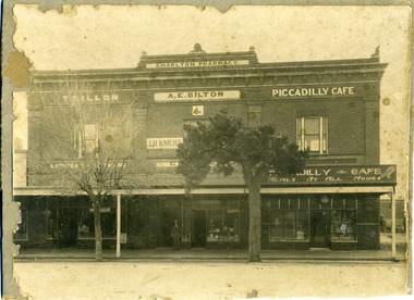 Sepia photo of Double storey brick building with 3 store fronts. One man standing in front.