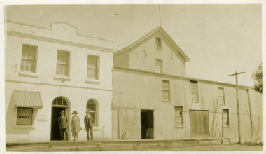 Two storey white brick building and corrugated building. Three people in front.