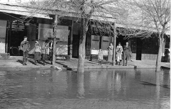 Flood waters in the main street. People are standing in front of shops
