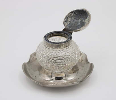 Glass inkwell with open silver lid, standing on silver dish stand.