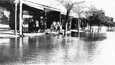 Flood water lap at front of shops in High Street, People are gathered under the veranda.