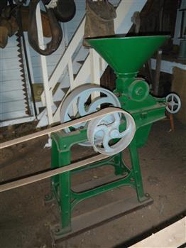 Green metal grain grinder with four legs that sit on two  wooden bases. It has two drive wheels painted blue.  