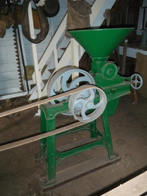 Green metal grain grinder with four legs that sit on two  wooden bases. It has two drive wheels painted blue.  