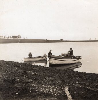 Black and white photograph of a whale boat and a dinghy with an outboard motor. Three men sit and stand in the two boats.