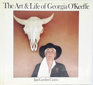 An older woman, her head covered by a long white scarf under a black hat, stands against an orange wall. Above, to her left, her hangs the skull of a steer. 