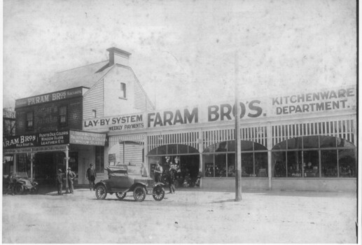Two storey weatherboard building and an adjoining long horizontal single storey building with large arched windows. The name 'Faram Bros' is prominent on both buildings. Several are outside the buildings in small groups and two early 20th century cars can be seen parked on the road.