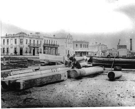 Three men sit on timber and prepared logs beside several small boats that have been pulled up onto the beach. Two-storey commercial buildings line the street in the background. 