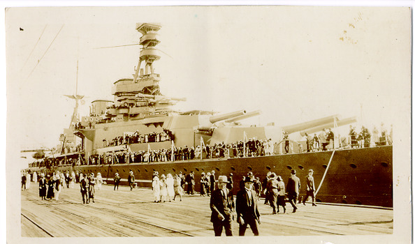 Many people are walking on a pier beside a large battleship. The crew are lined up on the decks of the ship.