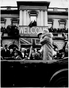 Black & white photo of  - Duke and Duchess of a couple standing in a car, passing a building with a balcony where a Welcome sign is hanging.   Groups of people can be seen at the back of the car and on the balcony of the building.