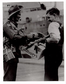 A man with a Cadbury's vendor tray slung around his shoulders sells chocolate to a second man in military uniform. The mid section of a ship can be seen in the background.