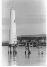 A lighthouse-like structure sitting in the sea on pylons with the remaining few metres of a dilapidated wooden walkway attached.