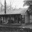Some people on a railway platform shelter from the rain under the corrugated iron roof of a modest wooden building. Four are sitting on a bench and one standing near the entrance, 