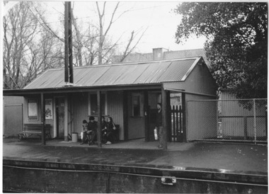 Some people on a railway platform shelter from the rain under the corrugated iron roof of a modest wooden building. Four are sitting on a bench and one standing near the entrance, 