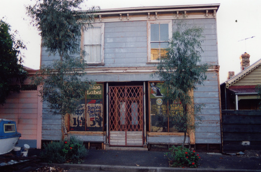 A two-storey double-fronted weatherboard building. Shop windows on the ground floor bear old grocery prices and advertisements.