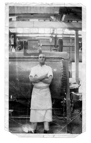 A man in white work clothes stands, arms folded, in front of a piece of machinery.