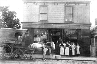 Four men in aprons and a lady in a black dress stand in front of a two-storey shop. A horse with a wagon stand in the roadway. The name 'W J Bellion' appears on the side of the wagon.