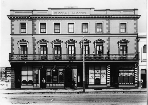 A large three-storey building with 'Royal Hotel' on the parapet. Four shops occupy the ground floor and some people are standing at the doorways of some of these. A man is standing sand a lady sitting on the first floor balcony.