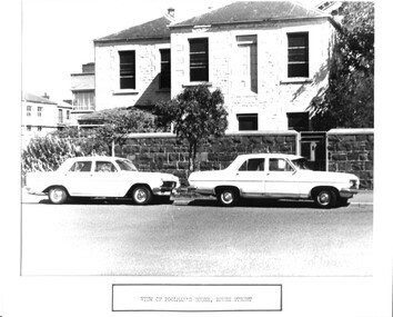 Two cars parked in front of a two storey stone house surrounded by a tall bluestone wall.