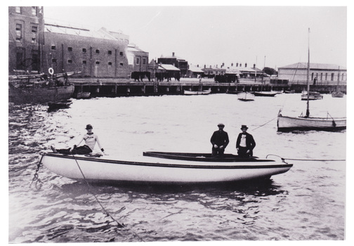 Two men stand in a boat moored in a small boat harbour while a third man sits of the prow. The background includes industrial buildings along the jetty and houses further back.