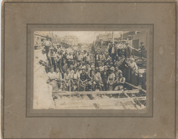 A large group of men in work clothes pose in a hole constructed in the middle of the road.