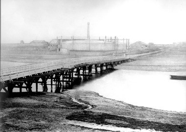 A wooden bridge crossing a body of water with gasworks in the background.